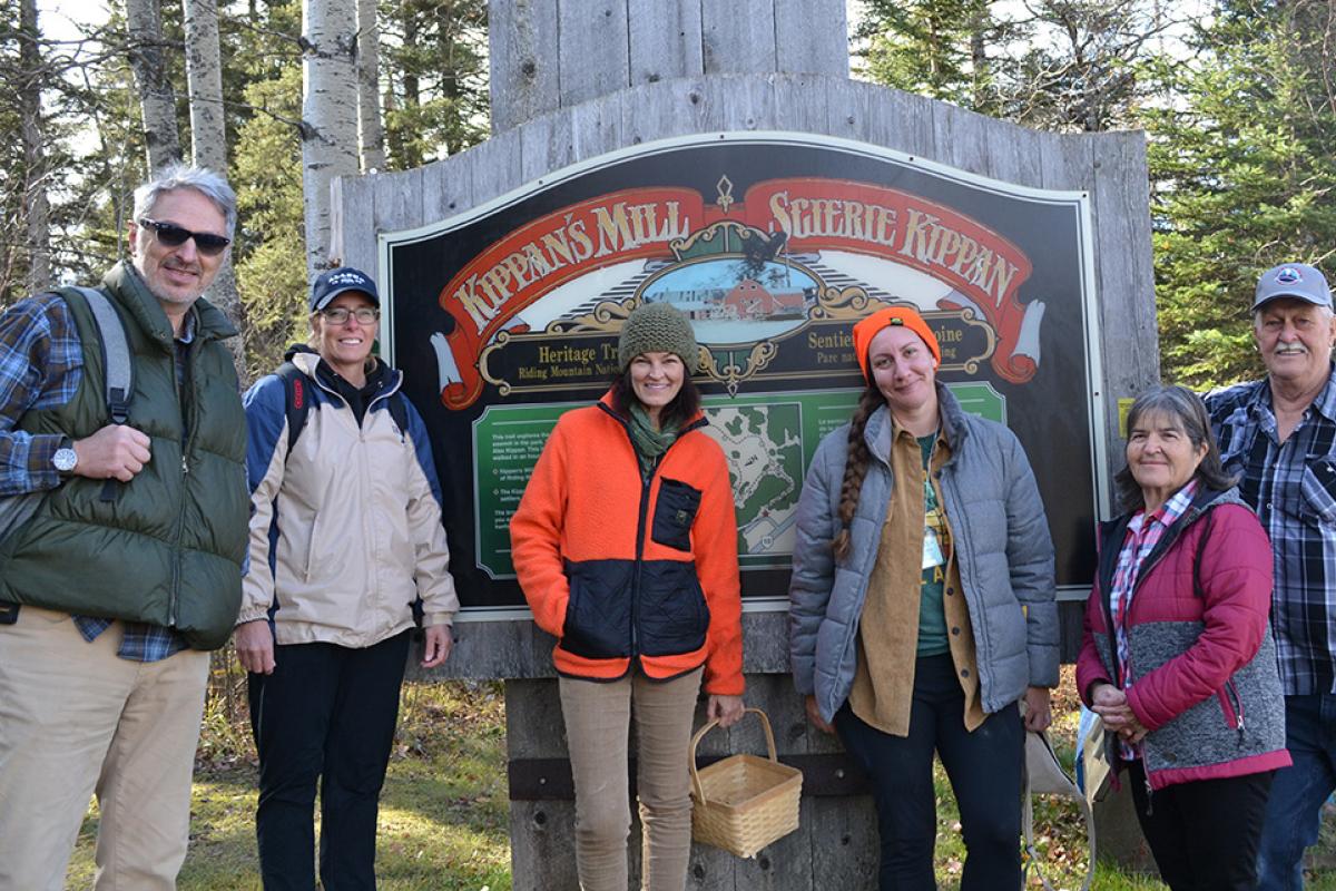 Six adults in hiking clothes stand in front of a trail head sign. 