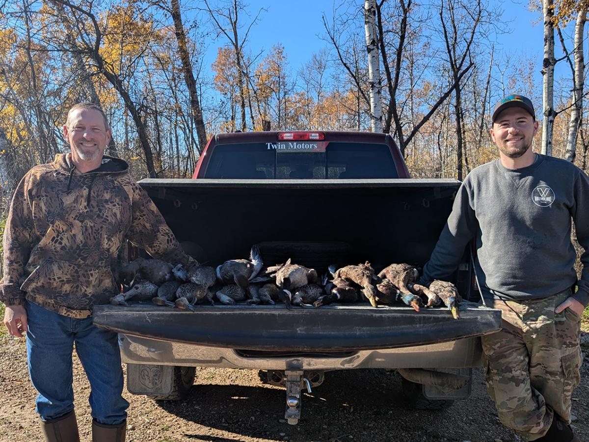 Two men stand on either side of a truck, with ducks from their recent hunt spread across the tailgate.
