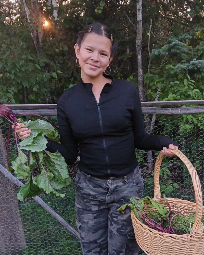 A young woman stands in a garden with forest behind. In one had she holds a basket full of vegetable and in the other hand she shows off a fresh beet.
